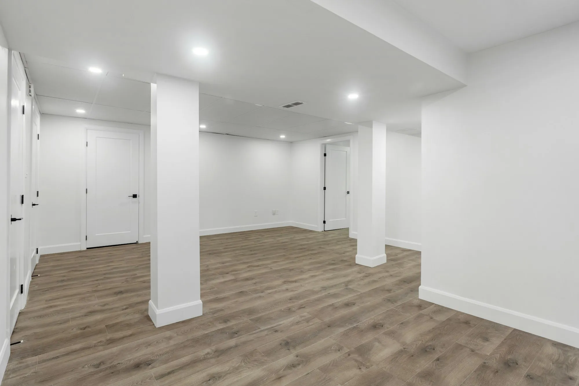 Finished basement with white walls, recessed lights, wrapped columns and wood-look flooring